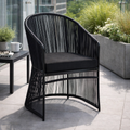 Black wicker chair with a cushion on a patio with plants and a table in the background.
