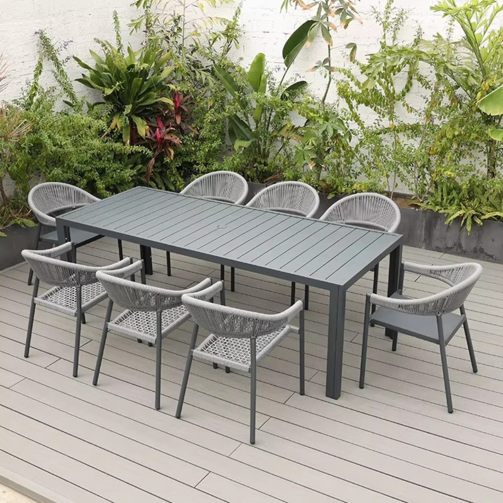 Outdoor patio set with a rectangular table and eight chairs on a wooden deck with plants in the background.
