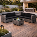 Outdoor patio set with black cushions on a wooden deck, surrounded by plants and a building.